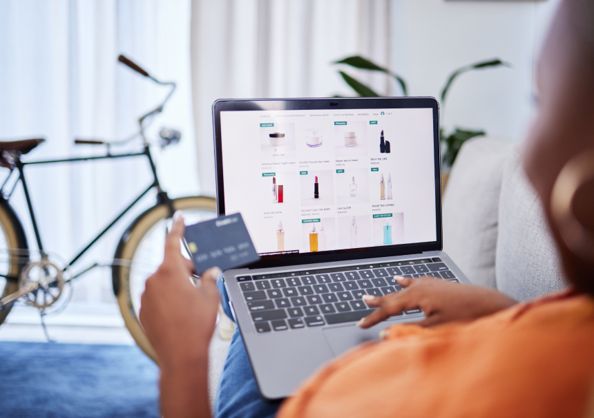 A woman shopping for cosmetics online on her laptop, holding a payment card. The screen displays a grid of personalized product recommendations driven by AI. A vintage bicycle is in the background.