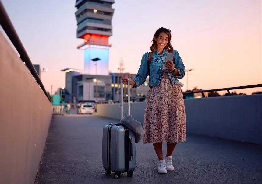 Smiling traveler with suitcase using a smartphone on an elevated pathway near a modern illuminated ATC tower at dusk.