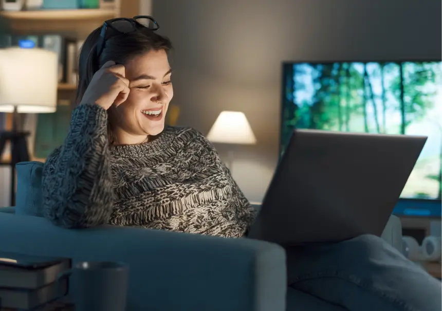 A happy woman engaging with a SaaS platform on her laptop and streaming media on the TV in a cozy evening living room.