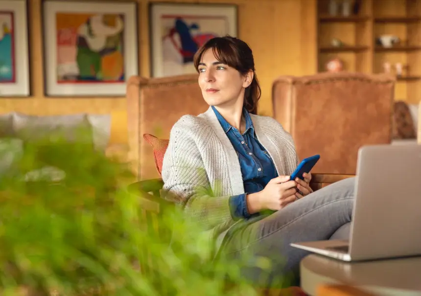 A woman sitting in an armchair holding a smartphone with a laptop nearby, working remotely during a break.