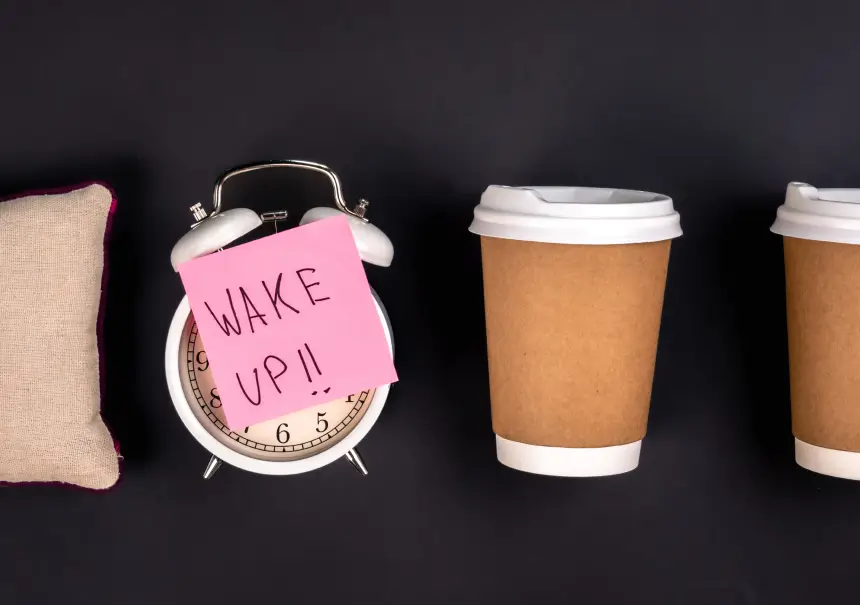 An alarm clock with a "Wake Up!!" sticky note next to a pillow and coffee cups on a dark background, illustrating an article about re-engaging dormant B2B leads