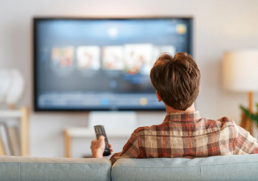 Rear view of a man sitting on a couch with a remote control, browsing personalized content recommendations on a smart TV streaming interface (VOD).