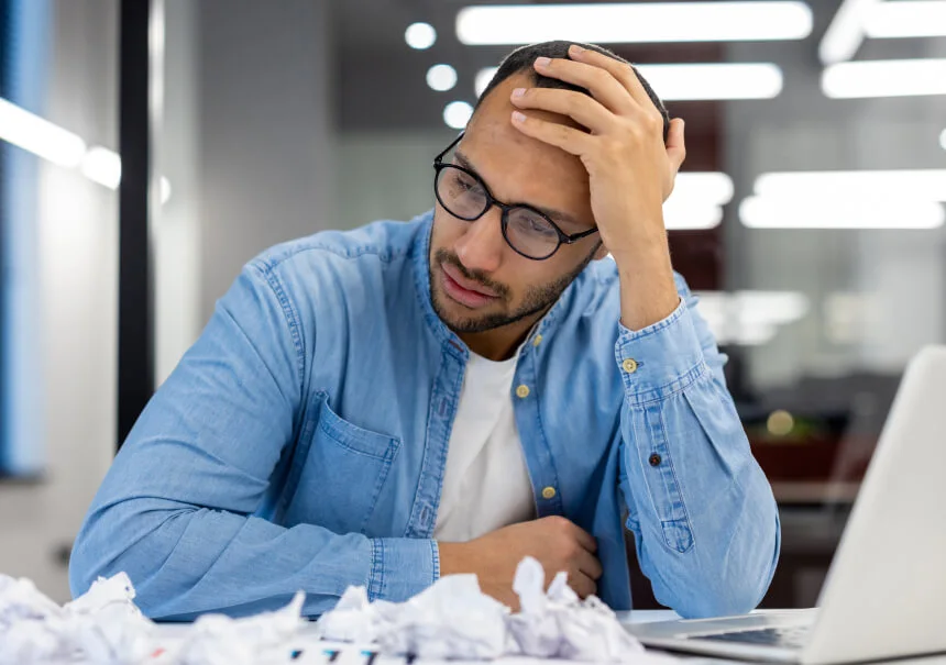 Stressed marketing operations manager holding his head in front of a laptop and crumpled papers in an office, depicting broken marketing automation workflows and funnel friction.