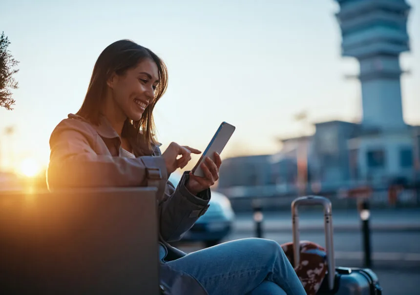 Smiling traveler checking a message on her smartphone at the airport at sunset, with a suitcase beside her