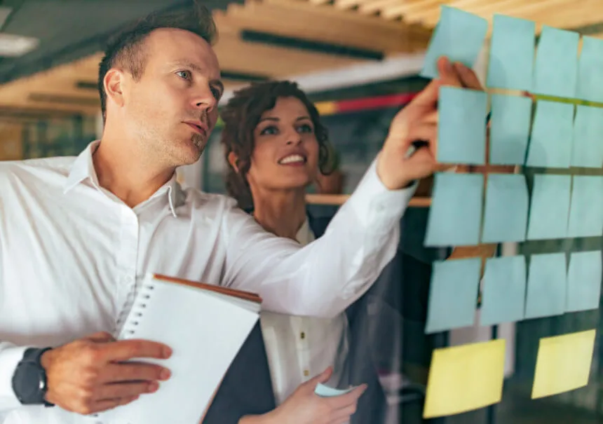 A marketing team designing a Customer Journey Map for marketing automation. A man and a woman placing sticky notes on a glass board to outline touchpoints, triggers, and automated workflows.
