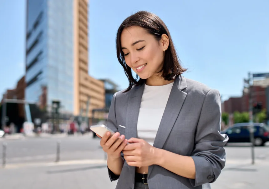 An e-commerce customer receiving a WhatsApp message from an online store. A smiling woman in a grey blazer uses her smartphone on a modern city street.