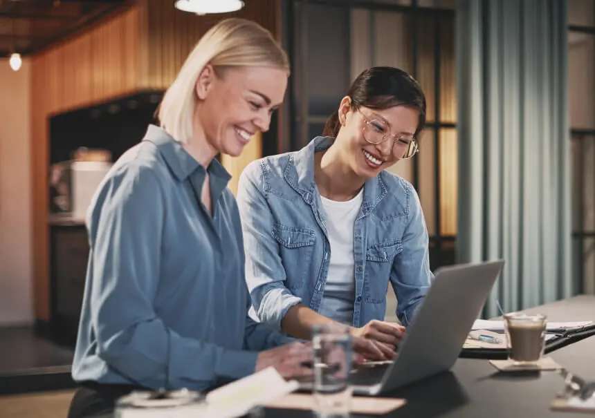 Two smiling marketing specialists designing a high-converting landing page using a no-code landing page builder. Women working together on a laptop in a modern marketing agency office.
