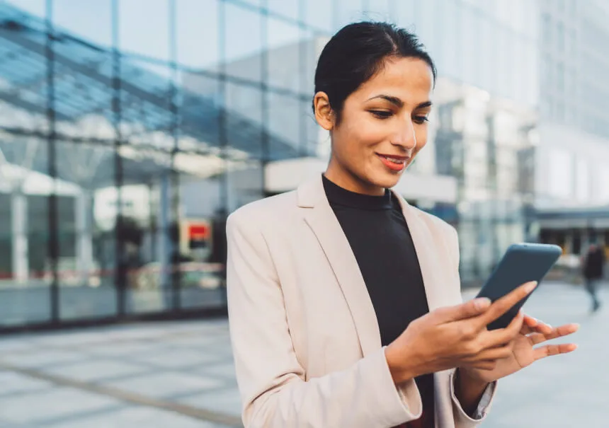 A new customer receiving a personalized welcome email on her smartphone. A smiling woman in a light blazer uses her phone in front of a modern office building, interacting with an automated customer onboarding workflow.
