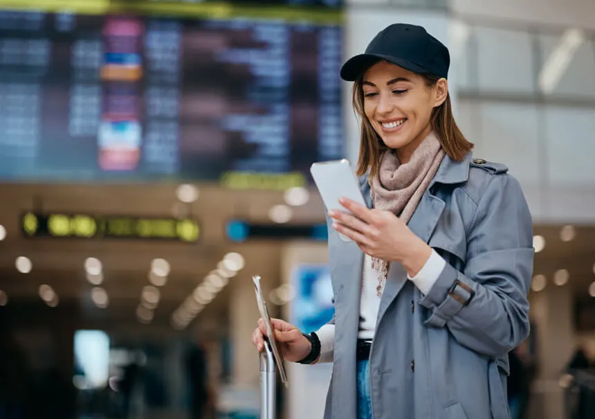 A smiling traveler with luggage at an airport reading an automated WhatsApp notification with booking details on her smartphone.