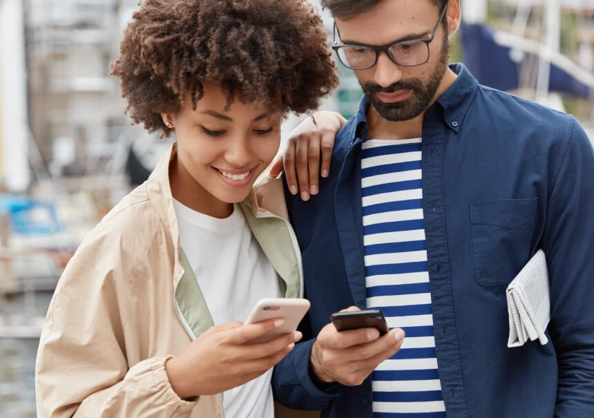 A smiling couple checking mobile app push notifications on their smartphones outdoors.