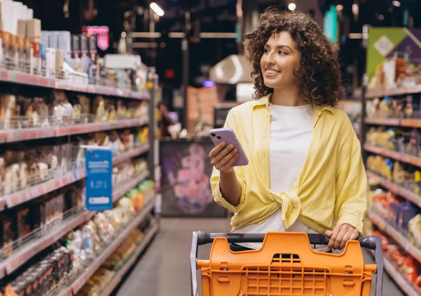 A smiling woman with a shopping cart holding a smartphone to check a WhatsApp marketing offer inside a grocery store.